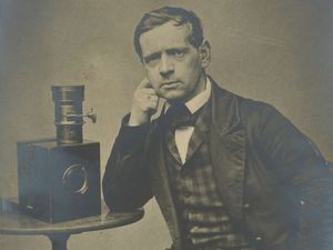 Selbstporträt von Charles Durheim, zeigt ihn sitzend und mit einem Arm auf einem Tischen abgestützt, auf welchem sich eine Kamera befindet.