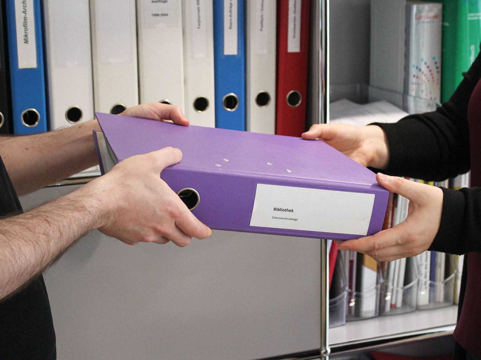 Eine Person übergibt einen Aktenordner an eine Mitarbeiterin der Burgerbibliothek.